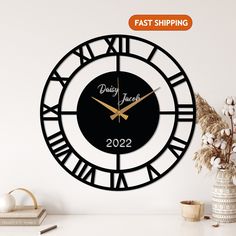 a black and gold clock on a white wall next to a vase with dried flowers