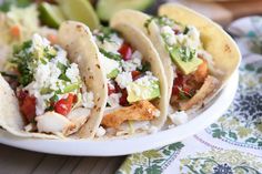 three fish tacos on a plate with avocado and feta in the background