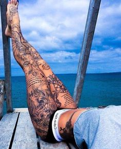 a man with tattoos on his legs sitting on a dock next to the ocean and looking at the water