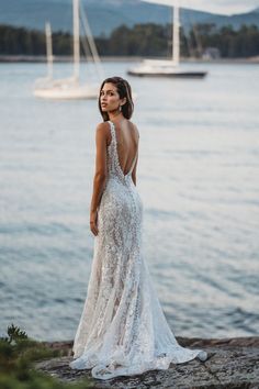 a woman in a wedding dress standing on the shore looking out at the water and yachts