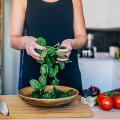 a woman is holding spinach leaves over a bowl on a cutting board next to tomatoes and other vegetables