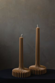 two tall candles sitting on top of a table next to each other in front of a gray wall