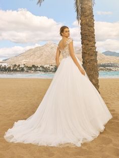 a woman in a wedding dress standing on the beach next to a palm tree and looking off into the distance