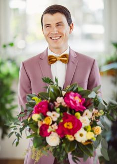 a man in a pink suit and bow tie holding a bouquet of flowers smiling at the camera