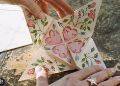 two hands are holding origami pieces with flowers on them and one hand is reaching for the paper