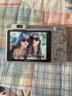 a digital camera sitting on top of a bed next to a plaid cloth covered blanket