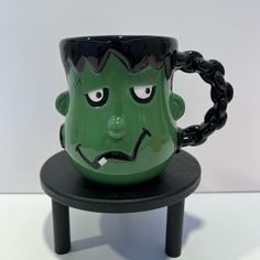 a green mug with a face on it sitting on a black table next to a white wall