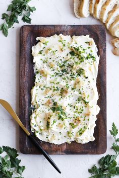 a wooden cutting board topped with mashed potatoes and parsley next to sliced bread