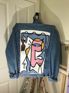 a blue jean jacket with an abstract painting on the front and back, hanging up against a white wall