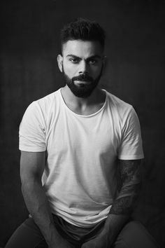 a man with a beard sitting in front of a black background wearing a white t - shirt