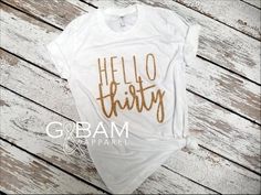 a white shirt with gold lettering that says hello thirty on the front and back, sitting on top of a wooden floor