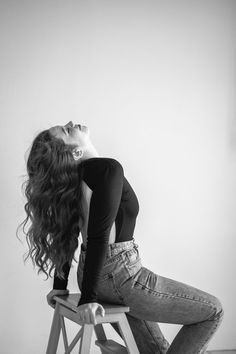 a woman sitting on top of a wooden chair next to a white wall with her hair blowing in the wind