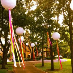 some white paper lanterns hanging from trees in front of a house with ribbons on them