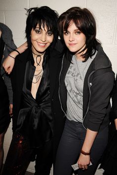 two women standing next to each other in front of a white wall and wearing black clothing