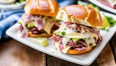 two sandwiches with meat, cheese and vegetables on a white plate sitting on a wooden table