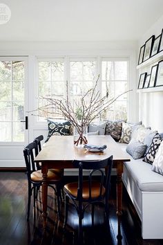 a dining room table surrounded by chairs in front of two windows with white walls and wooden flooring