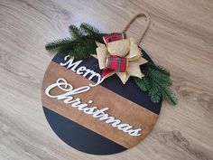 a merry christmas ornament hanging on a wooden board with pine branches and ribbon