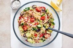 a glass bowl filled with couscous, tomatoes and cucumbers next to a lemon wedge