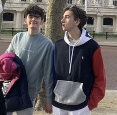 two young men standing next to each other in front of a tree on a sidewalk