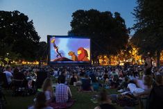 people sitting in the grass watching a movie on a large screen at night with bright lights