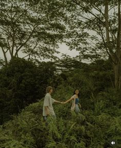 two people holding hands while walking through the grass in front of some trees and bushes