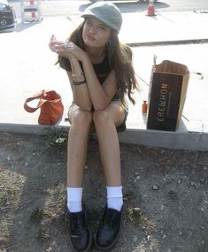 a young woman sitting on the ground with her hand under her chin, wearing black shoes and a hat