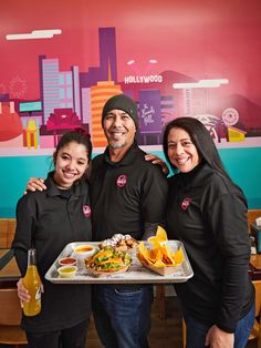 three people standing in front of a tray with food and drinks on it, smiling at the camera