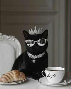 a black cat wearing glasses and a tiara sitting next to a coffee cup with a croissant on it