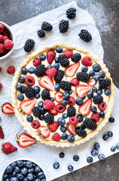 a pie topped with berries and blueberries on top of a white paper next to bowls of strawberries