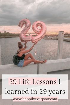 a woman sitting on the edge of a pier holding up a large pink number twenty