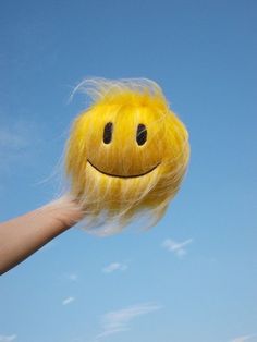 a hand holding up a yellow smiley face hair ball in the air with blue sky behind it