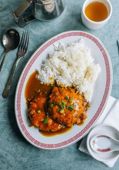 a white plate topped with meat covered in gravy next to rice and tea