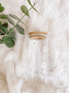 a glass jar with white flowers on it next to a green leafy plant in the background