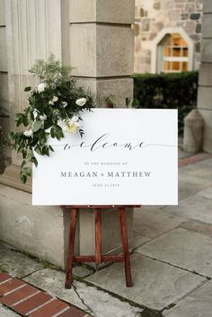 a welcome sign with flowers on it sitting in front of a stone building next to a wooden easel