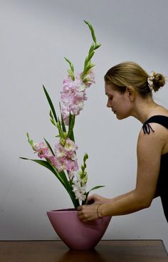 a woman arranging flowers in a pink bowl on a wooden table next to a white wall