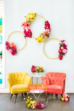 two chairs and a table with flowers hanging from the wall behind them in front of a white wall