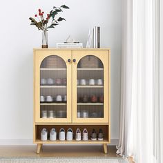 a wooden cabinet with glass doors next to a vase and flowers on the floor in front of a window