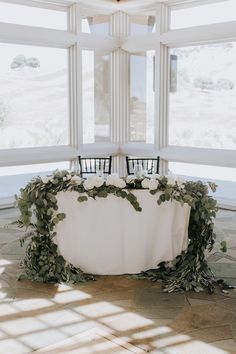 a table with white flowers and greenery on it in front of some large windows