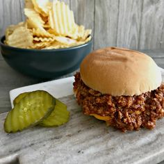 a sloppy joe sandwich next to a bowl of chips and pickle wedges on a marble tray