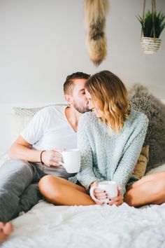 a man and woman sitting on top of a bed next to each other holding coffee cups