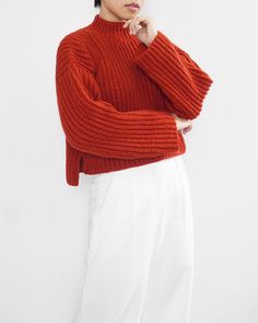 a woman in an orange sweater and white pants poses with her hands on her chin