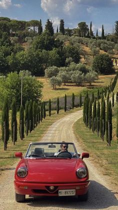 a red sports car driving down a dirt road with trees on both sides and a man in the driver's seat