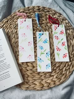 three bookmarks with watercolor flowers are sitting on a wicker basket next to an open book