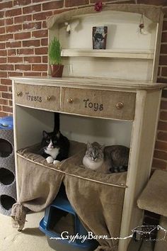 two cats laying on top of a cat bed under a desk with toys underneath it