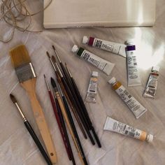 an assortment of paint and brushes laid out on a bed with a white bag in the background