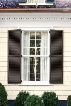 an open window with black shutters on a white brick building in front of green bushes