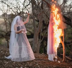 a woman in a wedding dress standing next to a fire with her hands raised up