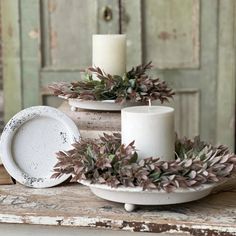 some white candles are sitting on a table next to other dishes and vases with greenery in them