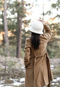 a woman standing in the woods wearing a coat and hat with her hands up above her head