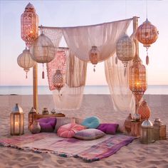 an outdoor seating area on the beach with lanterns hanging from it's ceiling and pillows